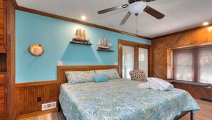 Un dormitorio con cama, paredes de madera, ventilador de techo y dos ventanas con persianas.