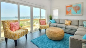 A living room with a view of the ocean and a pier, featuring a sofa, armchairs, and a round ottoman. - Oceanfront Monkeytyme in Oak Island: 5BR, 4BA, sleeps 13, perfect for families! (Oak Island)