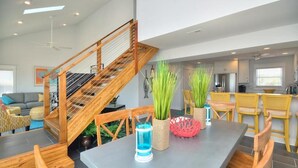 A spacious living area with a wooden staircase, a dining table with chairs, and a kitchen area in the background. - Oceanfront Monkeytyme in Oak Island: 5BR, 4BA, sleeps 13, perfect for families! (Oak Island)