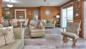 Ein geräumiges Wohnzimmer mit Holzboden, Deckenventilator, Essbereich mit weißen Stühlen, einem grünen Sofa und einem Holzschaukelstuhl.