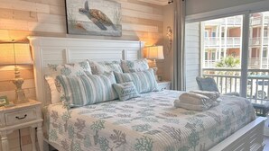 A bedroom with a white bed, striped pillows, a coral-patterned bedspread, a turtle painting above the bed, and a balcony with a view of buildings.