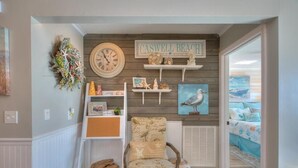 A room with a wooden wall, a clock, a 'CASWELL BEACH' sign, a chair, and a bed with a beach-themed bedspread.