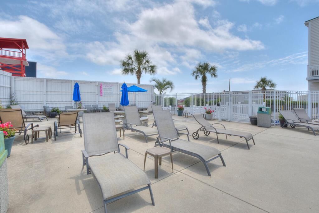 Une terrasse sur le toit, dotée de chaises longues et de parasols, entourée d’une clôture blanche et de palmiers.