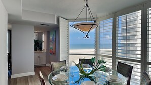 Uma sala de jantar com mesa e cadeiras de vidro, luminária pendente e vista para a praia através de grandes janelas.