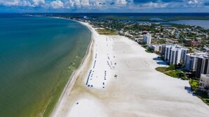 Vista aérea de uma área costeira com uma extensa praia de areia, uma área residencial densa com edifícios altos e um céu azul límpido.