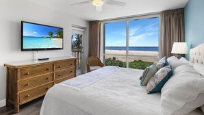 Quarto com cama, televisão, ventilador de teto e vista para a praia.