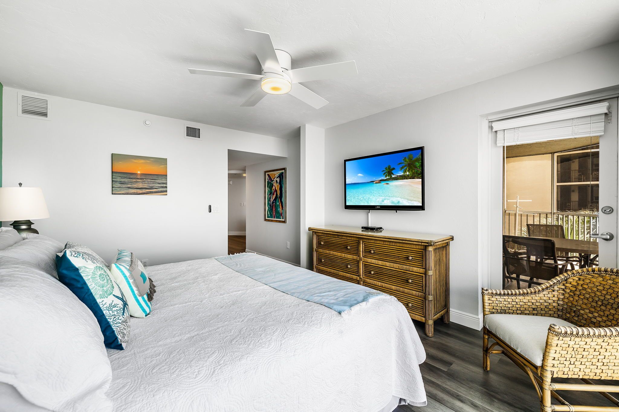 Un dormitorio con cama, televisión, una silla de ratán y vista a la playa a través de la ventana.