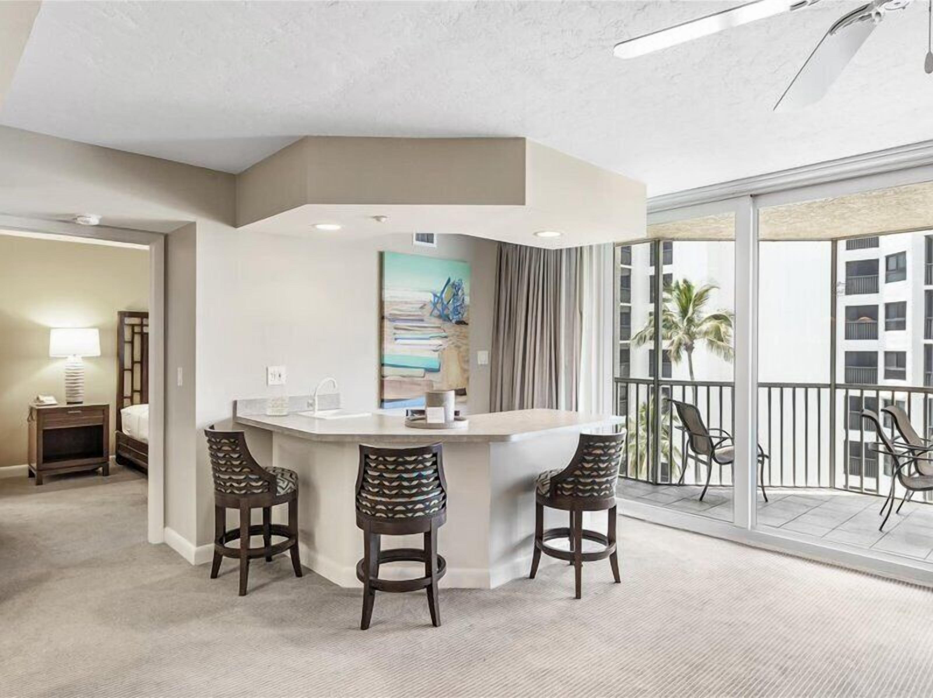 A hotel room with a bar area, two patterned chairs, a painting, and a balcony with chairs.