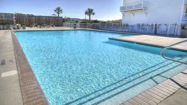 A clear swimming pool with a tiled deck, a wooden fence, and a building with a white exterior and a sign.