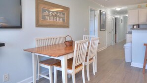 A dining area with a wooden table and white chairs, a framed picture on the wall, and a kitchen in the background. - Stay at Beach Pals in Oak Island—spacious condo with ocean views, sleeps 10! (Oak Island)
