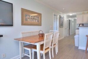 A dining area with a wooden table and white chairs, a framed picture on the wall, and a kitchen in the background.