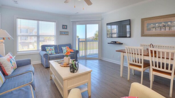 A spacious living room with a ceiling fan, a TV mounted on the wall, and a dining area with a wooden table and chairs. - Stay at Beach Pals in Oak Island—spacious condo with ocean views, sleeps 10! (Oak Island)