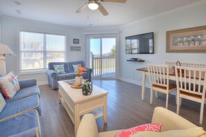 A spacious living room with a ceiling fan, a TV mounted on the wall, and a dining area with a wooden table and chairs. - Stay at Beach Pals in Oak Island—spacious condo with ocean views, sleeps 10! (Oak Island)
