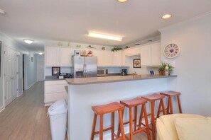 A modern kitchen with white cabinets, a stainless steel refrigerator, a microwave, and a coffee maker. There are wooden stools at the island counter.