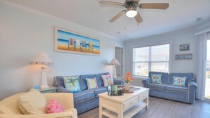 A living room with a blue sofa, a white coffee table, and a ceiling fan. - Stay at Beach Pals in Oak Island—spacious condo with ocean views, sleeps 10! (Oak Island)