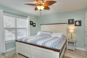 Quarto com cama, ventilador de teto, mesa de cabeceira com abajur e quadros nas paredes.