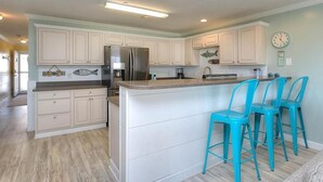 A kitchen with white cabinets, a stainless steel refrigerator, and a bar area with turquoise chairs. - Experience ocean views at Oak Island OW 3204, a spacious 3-bedroom condo ideal for 10 guests. (Oak Island)
