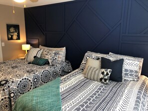 A hotel room with two beds, patterned bedding, a dark headboard, a wall-mounted lamp, and a framed picture.