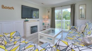 A living room with a sofa set, a fireplace, a television, and a balcony with a view of greenery. - Oceanview Condo OW 4203 in Oak Island: 2 bedrooms, sleeps 6, with stunning views! (Oak Island)