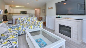 A living room with a sofa, coffee table, fireplace, TV, and kitchen in the background. - Oceanview Condo OW 4203 in Oak Island: 2 bedrooms, sleeps 6, with stunning views! (Oak Island)