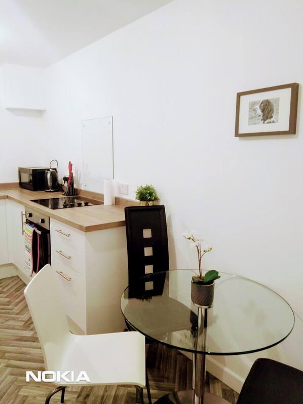 A modern kitchen with a microwave, kettle, and sink. There is a glass table with chairs and a small potted plant. A framed picture is on the wall.