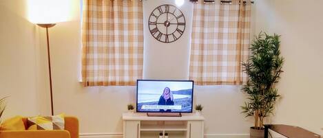 Uma sala de estar com televisão, relógio, sofá, abajur e planta.