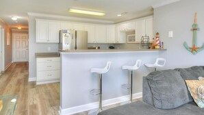 A modern kitchen with white cabinets, a central island with bar stools, and a nautical-themed wall decoration. - Enjoy ocean views in this spacious 3-bedroom condo on Oak Island! (Oak Island)