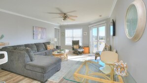 A spacious living room with a grey sofa, a glass coffee table, and a ceiling fan. - Enjoy ocean views in this spacious 3-bedroom condo on Oak Island! (Oak Island)