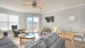 A spacious living area with a ceiling fan, a sofa, a coffee table, a TV mounted on the wall, and a dining table with chairs. - Enjoy ocean views in this spacious 3-bedroom condo on Oak Island! (Oak Island)