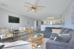 Ein modernes Wohnzimmer mit einem grauen Sofa, einem Glastisch und einem Deckenventilator. Es gibt eine Essecke mit Tisch und Stühlen, und im Hintergrund befindet sich eine Küche.