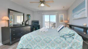 A bedroom with a bed, a ceiling fan, a dresser, a TV, and a large window. - Enjoy ocean views in this spacious 3-bedroom condo on Oak Island! (Oak Island)