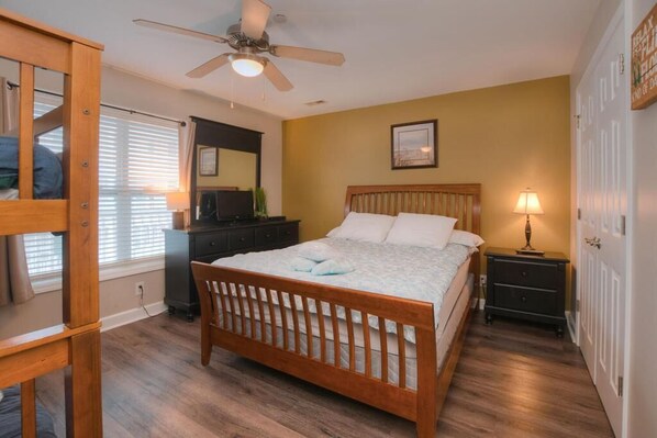 A bedroom with a wooden bed, a ceiling fan, a dresser, and a nightstand with a lamp. - Relax in this oceanview condo in Oak Island, featuring 2 bedrooms for 6 guests. (Oak Island)