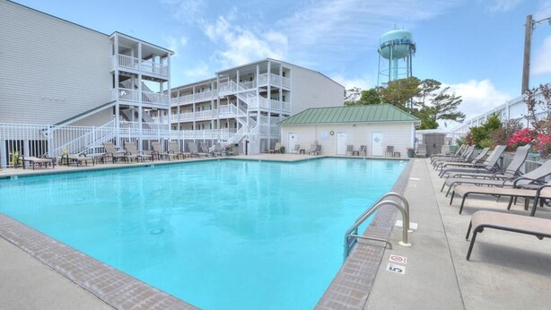A swimming pool with a clear blue water, a white building with multiple balconies, and a water tower in the background. - Relax in this oceanview condo in Oak Island, featuring 2 bedrooms for 6 guests. (Oak Island)