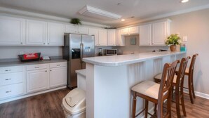 A kitchen with white cabinets, a stainless steel refrigerator, a microwave, and a small island with bar stools. - Relax in this oceanview condo in Oak Island, featuring 2 bedrooms for 6 guests. (Oak Island)