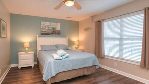 A bedroom with a bed, two bedside tables, a ceiling fan, a window with blinds, and a wall-mounted artwork. - Relax in this oceanview condo in Oak Island, featuring 2 bedrooms for 6 guests. (Oak Island)