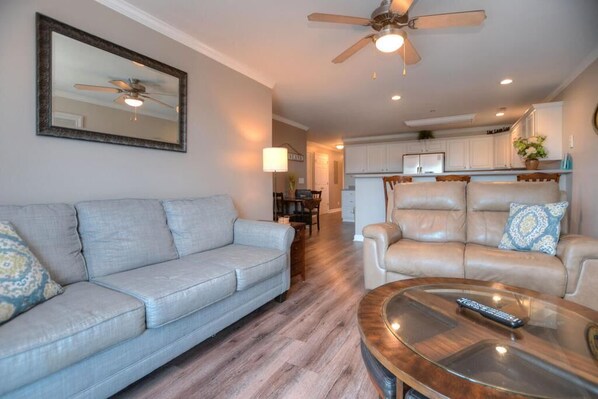 Een woonkamer met een grijze bank, een beige fauteuil en een glazen salontafel met een afstandsbediening.