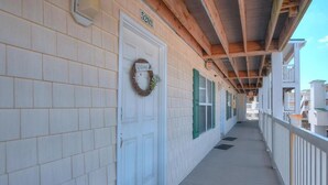 A covered wooden porch with a white door marked '5203', a welcome wreath, and a view of the adjacent buildings. - Relax in this oceanview condo in Oak Island, featuring 2 bedrooms for 6 guests. (Oak Island)