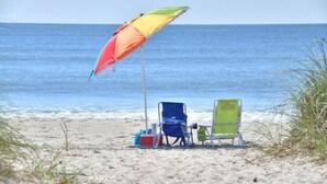 A beach scene with a colorful umbrella, two beach chairs, and a cooler on the sand. - Spacious oceanview condo in Oak Island, perfect for up to 9 guests with 3 bedrooms. (Oak Island)