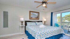 A bedroom with a bed, two bedside tables with lamps, a ceiling fan, a window with a view, and a blue chair outside. - Explore Oak Island from OW 6204-Anchors Away, a 3BR oceanview condo for 8. (Oak Island)