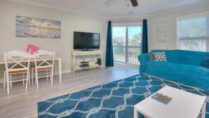A living room with a blue sofa, a white coffee table, a TV on a white stand, a ceiling fan, and a painting of a beach scene. - Explore Oak Island from OW 6204-Anchors Away, a 3BR oceanview condo for 8. (Oak Island)