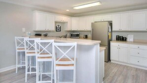 A modern kitchen with white cabinetry, a central island with bar stools, a refrigerator, and a microwave. - Explore Oak Island from OW 6204-Anchors Away, a 3BR oceanview condo for 8. (Oak Island)