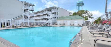 Una piscina con agua cristalina azul, un edificio blanco con varios balcones y una torre de agua al fondo.