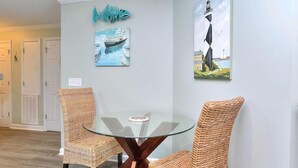 Un coin repas avec des chaises en osier et une table en verre, agrémenté d’œuvres d’art représentant un poisson et un phare sur les murs.