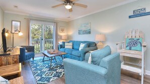 Un salon avec un canapé bleu, une table basse en bois et un ventilateur de plafond. Une grande fenêtre offre une vue sur les arbres, et il y a un balcon.
