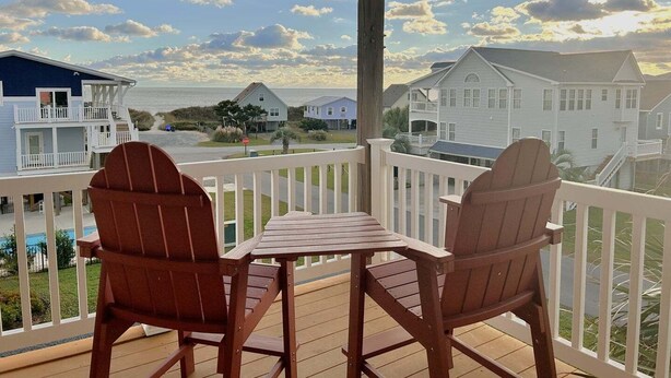 A wooden deck with two Adirondack chairs and a small table, overlooking a residential area with houses and a view of the ocean.