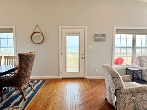 A room with a wooden floor, a dining table with chairs, a door leading to a balcony, and two windows with a view of the beach. - Experience oceanfront luxury at Pirate's View in Oak Island—perfect for 16 guests! (Oak Island)