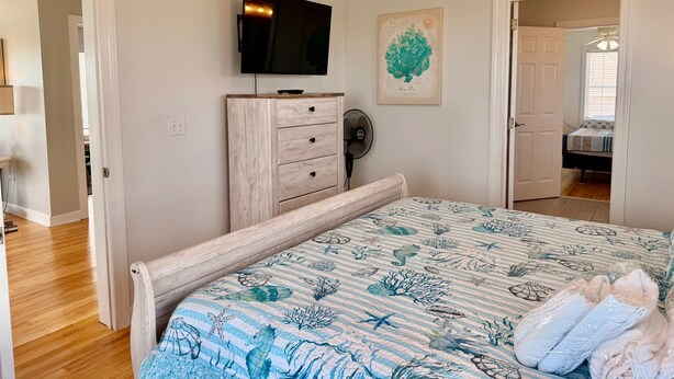 A bedroom with a bed, a dresser, a television, a fan, and a door leading to another room. - Experience oceanfront luxury at Pirate's View in Oak Island—perfect for 16 guests! (Oak Island)