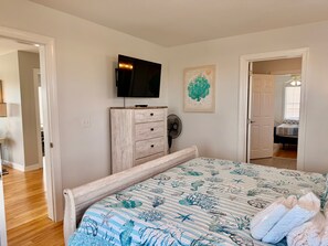 Une chambre à coucher comprenant un lit, une commode, un téléviseur, un ventilateur et une porte donnant sur une autre pièce.