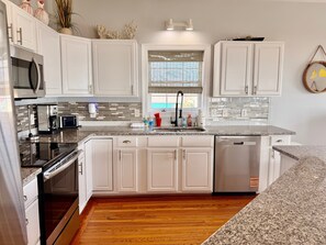 A modern kitchen with white cabinets, a granite countertop, stainless steel appliances, and a wooden floor. - Experience oceanfront luxury at Pirate's View in Oak Island—perfect for 16 guests! (Oak Island)