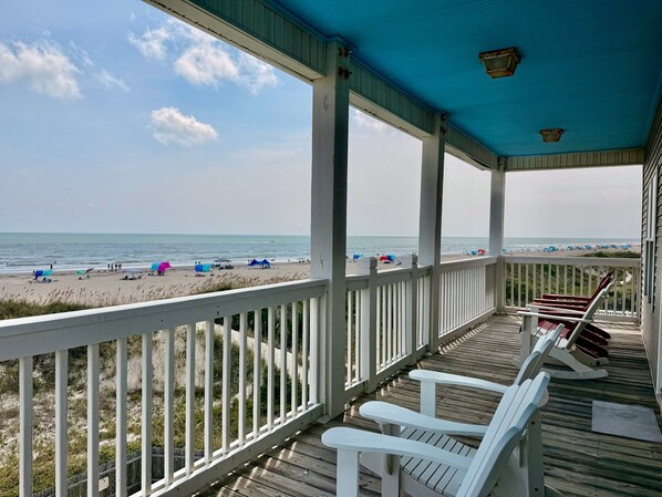 A covered deck with a view of the beach, white railings, and wooden flooring.